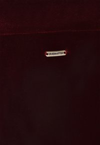Burgundy-mato, joka on pehmeä tekstuuriltaan; siinä on metallilappu, jossa lukee "DREIMASTER", kiinnitettynä kahdella pienellä punaisella langalla.