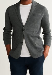 Šedý pletený blazer s klopovým límcem, dvouknoflíkovým zapínáním, bočními kapsami a strukturovaným povrchem, doplněný bílým tričkem a tmavými kalhotami.
