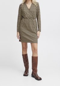  mekko, jossa on pitkät hihat ja V-pääntie, ruskeavalkoinen ruutukuosi, vyötärö kireällä, yhdistettynä polven yli ulottuviin ruskeisiin nahkasaappaisiin.