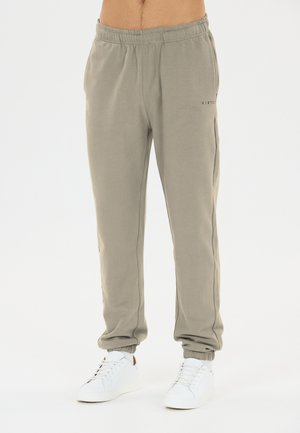 Homme portant un pantalon de jogging gris clair avec taille et poignets élastiques, associé à des baskets blanches, debout devant un fond uni et clair.