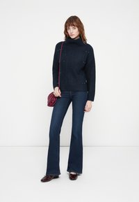 Pull en maille marine à col roulé avec motif texturé et perles décoratives, associé à un jean en denim noir évasé et un sac à main bordeaux.