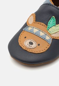 Chaussure en cuir navy avec un motif d'ours. Présente des accents beige et bleu clair, des détails brodés et une semelle en daim brun doux.