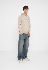 Beige hooded sweatshirt with a front pocket, featuring blue logo. Worn with loose-fitting, faded blue jeans and white sneakers.