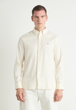 Cream long-sleeve button-up shirt with a collar, subtle texture, and a small logo on the chest. Paired with light-colored pants.