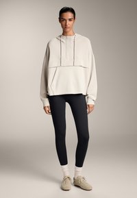 Hanorac oversized de culoare bej deschis, cu șnururi și buzunar frontal, purtat cu colanți negri și încălțăminte deschisă. Design simplu, modern.