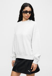 Γυναίκα με μακριά σκούρα μαλλιά που φοράει μαύρα γυαλιά ηλίου, φαρδιά ανοιχτογκρι φούτερ και μαύρη φούστα Puma μπροστά σε απλό φόντο.