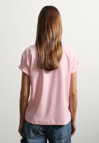 Top de manga corta, de color rosa claro, con un corte relajado y puños enrollados. Material suave y dobladillo redondeado, combinado con pantalones cortos de mezclilla.