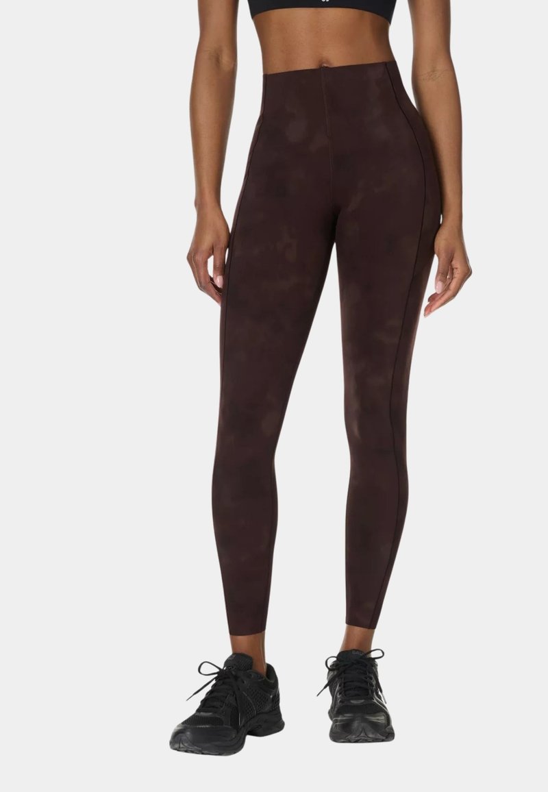 Woman wearing high-waisted dark brown leggings with subtle pattern and black athletic shoes, standing against a plain background.