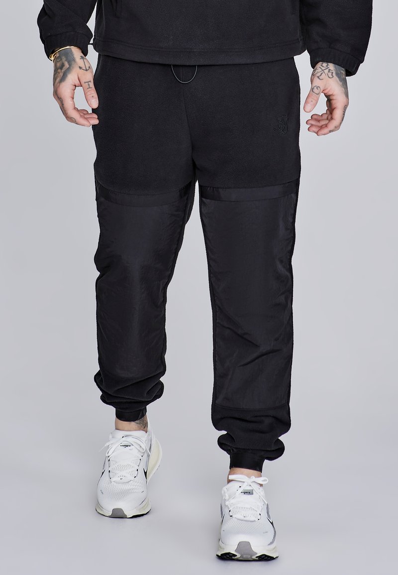 Čierne jogger nohavice s kombináciou mäkkého fleece a hladkých nylonových panelov, so elastickým pásom a manžetami, doplnené o detail loga.