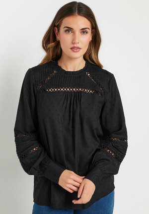 Blouse noire à manches longues avec tissu texturé, accents plissés, inserts en dentelle aux épaules et col rond.