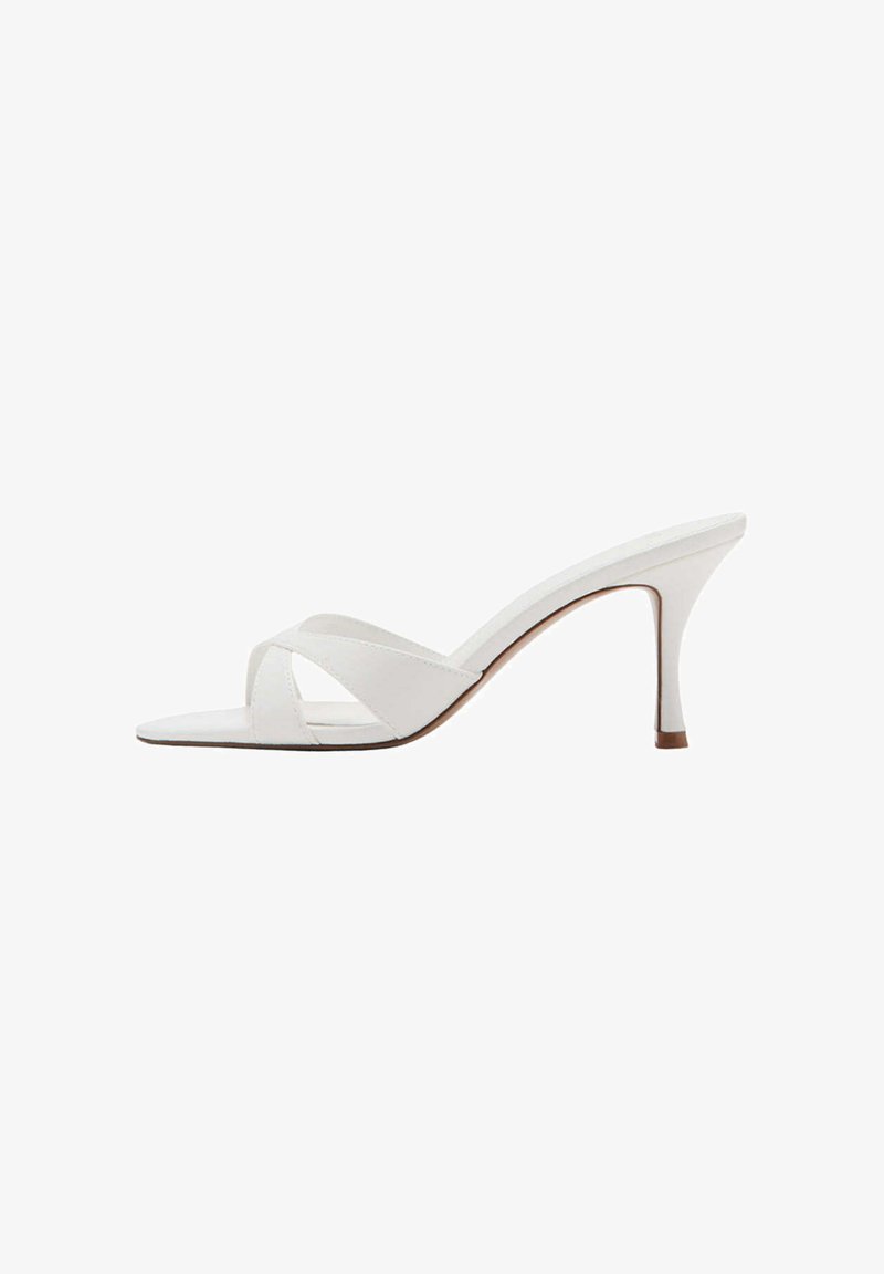 Mule blanche à bout ouvert avec des brides croisées et un talon stiletto fin de hauteur moyenne, montrée de profil sur un fond blanc.