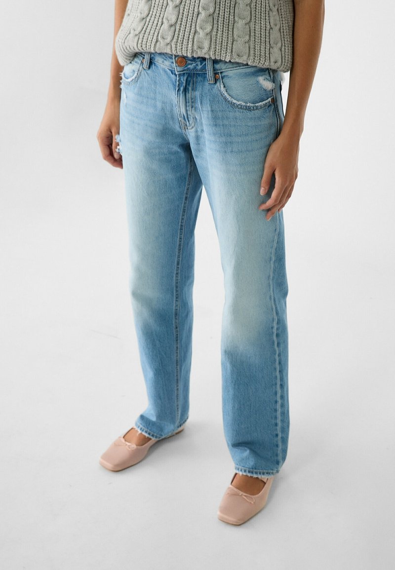 Jeans rectos de un azul claro con un lavado desteñido, dobladillo deshilachado y diseño clásico de cinco bolsillos, combinados con un par de zapatos planos beige y una blusa de punto texturizada.