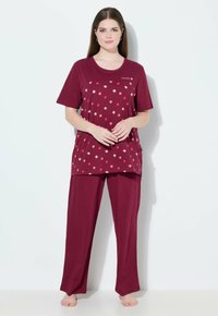 Frau steht barfuß und trägt ein weinrotes Pajama-Set mit floralem Muster auf der Oberseite und dem Text "SWEET" in der Nähe des Halsausschnitts.