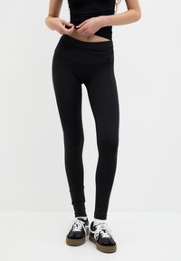 Fekete leggings, magas derékkal, sima textúrával, testhezálló kialakítással és enyhe fényességgel, fekete-fehér sportcipőkkel párosítva.