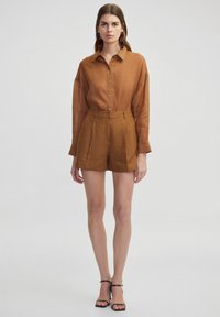 Brown linen button-up shirt with long sleeves paired with knee-length brown pleated shorts and black heeled sandals.