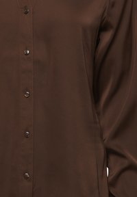 Blusa marrone con bottoni, realizzata in tessuto morbido, caratterizzata da una scollatura a V e sei bottoni abbinati sulla parte anteriore, con maniche lunghe.