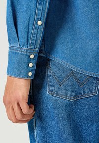 Manica di camicia in denim con tre bottoni a pressione bianchi, caratterizzata da un tessuto blu, una texture liscia e un motivo di cucitura a zigzag sulla tasca.