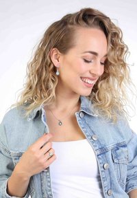 Druppelvormige turquoise oorbellen en een bijpassende hanger aan een fijne zilveren ketting. Het model draagt een licht denimjasje over een witte top.