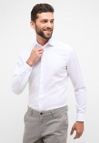 Camicia bianca a maniche lunghe con colletto classico, abbinata a pantaloni grigi, caratterizzati da una texture liscia e una vestibilità standard.