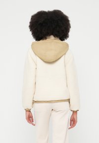 Chaqueta de forro polar en color crema con capucha color marrón claro. Presenta puños elásticos y dobladillo con cordón ajustable. Textura suave, diseño minimalista y ajuste casual.