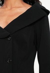 Melna garām piedurknēm topi ar platu asimetrisku atloku un divām melnām pogām priekšpusē, kas atklāj augšējo krūtīs un plecu zonu.