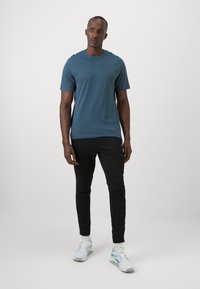 Teal t-skjorte med korte ermer og rund hals, kombinert med svarte slim-fit bukser og hvite joggesko med grønne og blå detaljer.