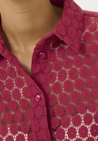 Krajková košile v tmavě růžové barvě s květinovým vzorem, s kulatými otvory a růžovými knoflíky na límci. Detailní textura je viditelná.