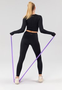Schwarzes Langarmshirt und hoch taillierte Leggings, beige Turnschuhe tragend, ein lila Widerstandsband haltend, mit glatter, eng anliegender Textur.