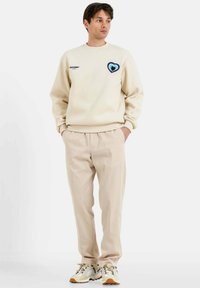Sudadera beige con un gráfico de corazón en azul y verde, combinada con pantalones beige y zapatillas blancas con suelas marrones, fondo liso.