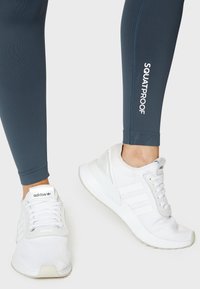 Dunkelblaue Leggings mit dem weißen Schriftzug "SQUATPROOF", kombiniert mit weißen Adidas-Sneakern, die über drei Streifen und ein strukturiertes Material verfügen.