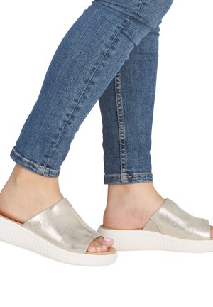 Feet wearing open-toe silver slip-on sandals with thick white soles and blue jeans rolled at the ankle, isolated on white background.