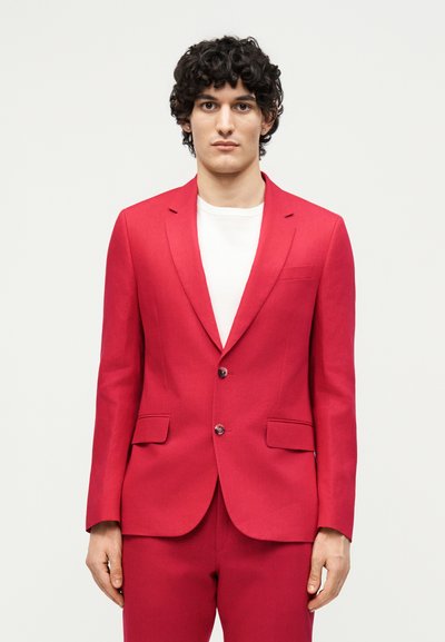 Άνδρας με σγουρά σκούρα μαλλιά που φορά φωτεινό κόκκινο σακάκι με δύο κουμπιά και αντίστοιχο παντελόνι πάνω από λευκό πουκάμισο, στέκεται μπροστά σε απλό φόντο.