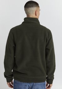 Pullover in pile verde scuro con colletto alto e polsini a coste, caratterizzato da una discreta patch con logo sul retro e una superficie liscia.