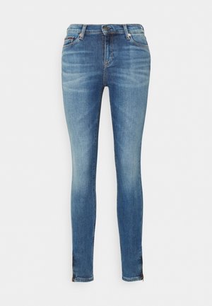 Jean skinny bleu avec bouton frontal, passants de ceinture et détails délavés sur les cuisses et les genoux, présenté sur fond blanc.