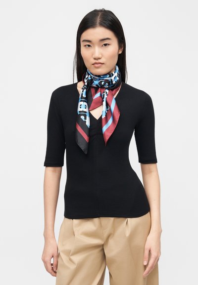 Jeune femme aux cheveux bruns et lisses portant un haut noir à manches courtes, un pantalon beige et un foulard en soie coloré noué autour du cou.