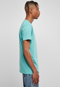 Tricou din bumbac turquoise, cu mâneci scurte și o croială normală, expus pe un model masculin văzut din lateral, pe un fundal neutru.