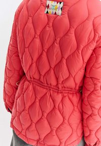 Prošívaná červená bunda s diamantovým vzorem šití a barevnou značkou Naoko na horní části zad, v pase stažená.