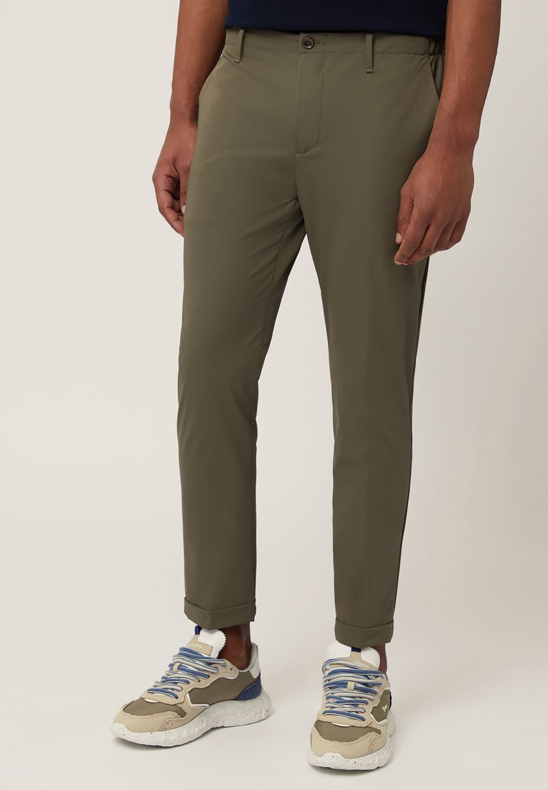 Pantalons fuselés vert olive en tissu léger, dotés d'une fermeture éclair et d'un bouton, avec des ourlets retroussés. Portés avec des baskets beiges et bleues.