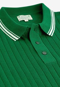Polo en maille côtelée verte avec col rayé blanc et deux boutons noirs sur la patte de boutonnage, étiquette indiquant la marque et la taille M.