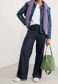 Kolorowa, pasiasta kurtka, biała koszulka, spodnie z wysokim stanem w ciemnym dżinsie o szerokich nogawkach, białe sneakersy, zielona torba z sznurkiem i uchwytami.