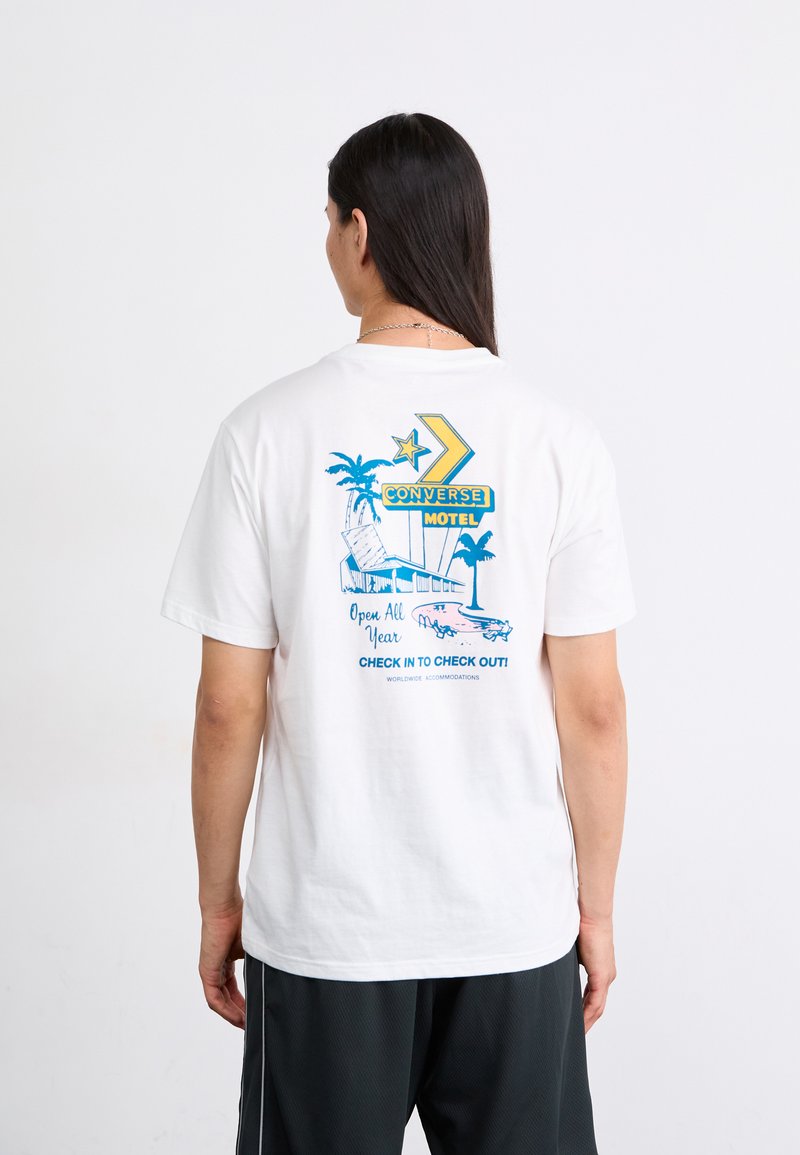 Hvid bomulds t-shirt med et blåt og gult retro motel grafik på ryggen, inklusive palmer og en illustration af en vintage bil.