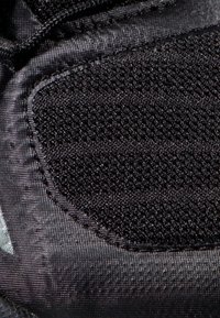 Tessuto nero testurizzato con sezioni in rete e lisce. Il materiale sembra duraturo con accenti cuciti e un dettaglio di logo riconoscibile.