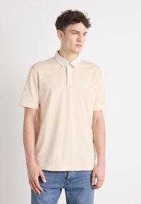 Beige kortärmad pikétröja med krage och en liten logotypdetalj på bröstet. Tillverkad av mjukt och slätt tyg.