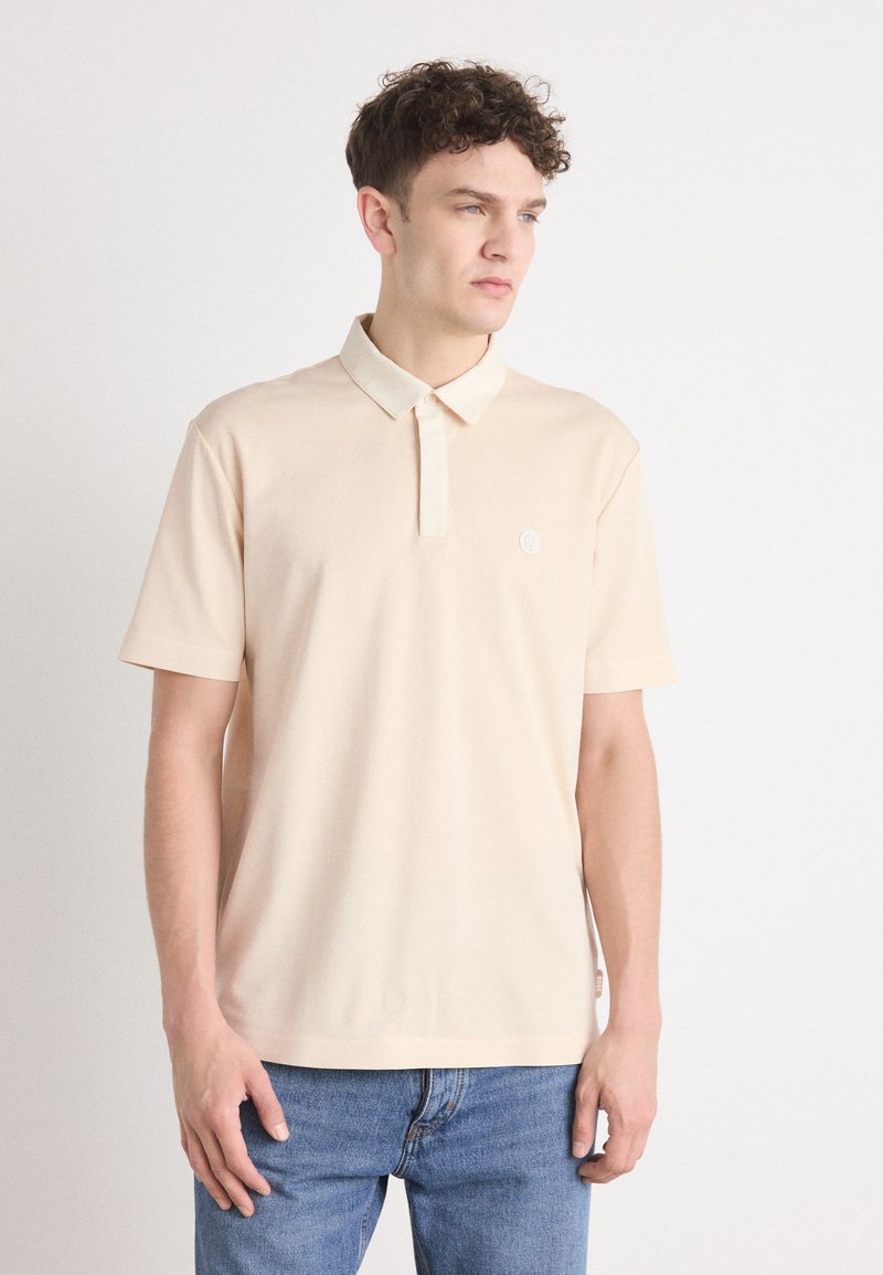 Beige kortärmad pikétröja med krage och en liten logotypdetalj på bröstet. Tillverkad av mjukt och slätt tyg.