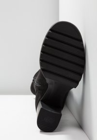 Bottine noire en daim texturé, avec une semelle en caoutchouc proéminente à rainures et un léger talon. Détail de fermeture éclair visible.