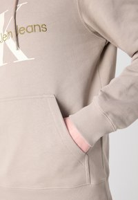 Hoodie beige con tasca a marsupio, polsini a costine e branding bianco con accenti verdi. Realizzato in morbido materiale misto cotone.