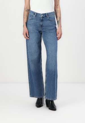 Model iført højtsiddende, lige blå jeans kombineret med sorte støvler med hæl og en hvid ærmeløs top, står på hvidt gulv.