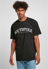 Juodas medvilnės marškinėliai su vingiuotu baltu „SOUTHPole ORIGINALS“ logotipu, trumpomis rankovėmis ir laisvu siluetu.