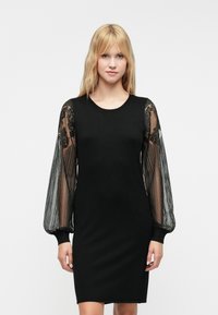 Juoda suknelė su prigludusia siluetu, turinti permatomas pūstines rankoves, puošnias juodu nėriniu ir raukšlėtomis apykaklėmis.