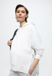 Ανοιχτό γκρι φούτερ με κουκούλα, άνετη εφαρμογή, πλαϊνές χαραγές και ριμπ μανσέτες. Φοριέται με μια μαύρη τσάντα ώμου. Το φόντο είναι απλό λευκό.